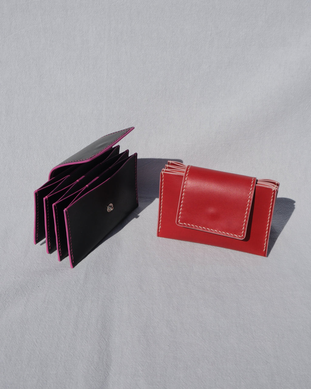 Two leather card holders on a light gray fabric background: one black with bright pink lining shown open, and one red with white stitching shown closed.